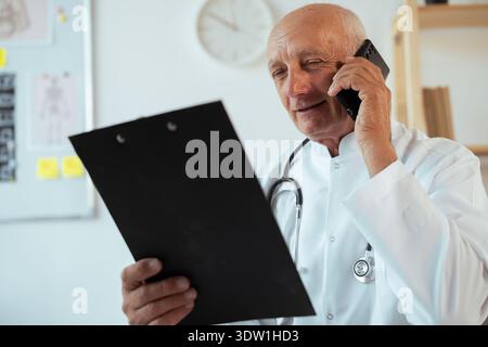 Senior man doctor with stethoscope making phone call, talking to patient scheduling appointments and taking notes Stock Photo