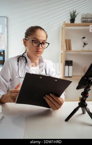 doctor wearing eyeglasses and white coat using phone on tripod, consulting patient online and taking notes. therapist making videocall in her medical Stock Photo