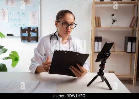 female doctor wearing eyeglasses and white coat using phone on tripod, consulting patient online and taking notes. therapist making videocall in her Stock Photo