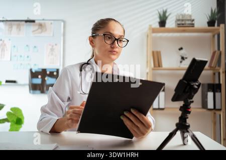 female doctor wearing eyeglasses and white coat using phone on tripod, consulting patient online and taking notes. therapist making videocall in her Stock Photo