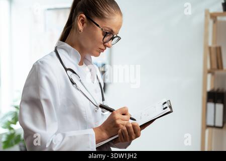 Young female doctor wear eyeglasses filling paper medical records. General Practitioner doing paperwork at workplace, writing notes and preparing Stock Photo
