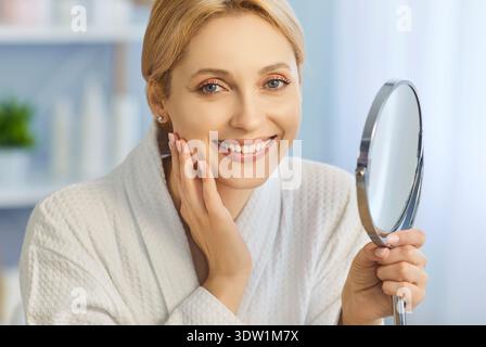 Attractive beautiful woman wearing bathrobe, smiling happily touching her face and holding mirror Stock Photo