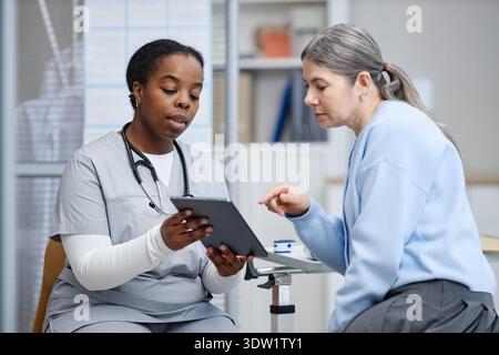 Young Adult Black Woman Doctor Showing Tablet to Middle Aged Caucasian Woman Stock Photo