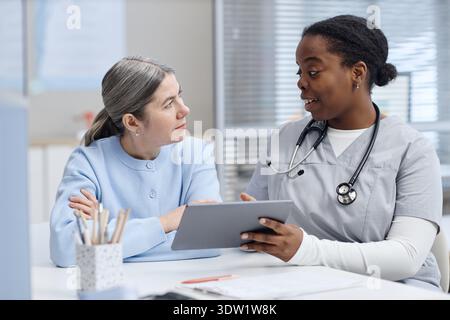 Mature Caucasian Woman Engaging with Black Female Nurse Using Digital Tablet Stock Photo