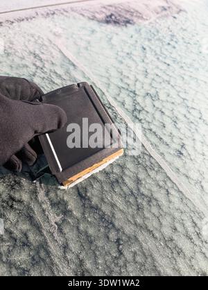 Close-up of a hand in a black glove scraping thick ice and frost from a car windshield with a plastic scraper. Stock Photo