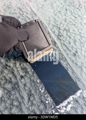 Close up of a hand in black glove using a plastic scraper to remove thick ice from a frozen car windshield. Stock Photo