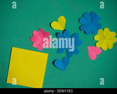 Yellow sticky notepad surrounded by paper flowers green background Stock Photo