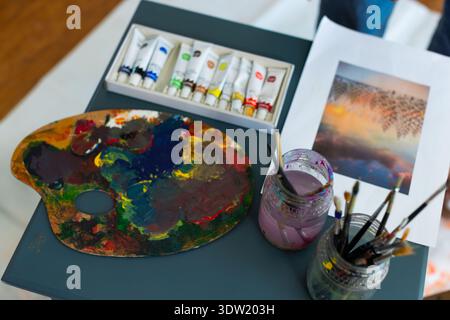 Artist palette is resting on gray-blue table with paint tubes, jars of brushes and reference print Stock Photo