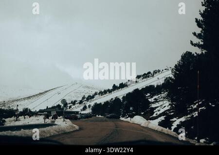 An empty road to a snowy mountain ski resort on a cloudy winter day. Scenic view of a roadway winding through a snowy mountain landscape under a grey Stock Photo