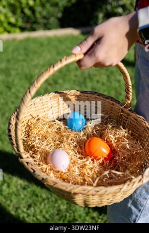 Female wearing smartwatch and denim pants holding wicker basket with three colorful eggs on lawn Stock Photo