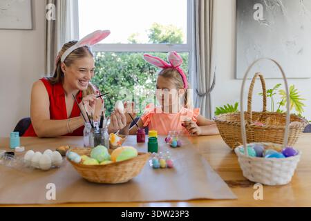 Adult woman and child painting eggs at wooden dining table, wearing bunny ears with paint jars Stock Photo