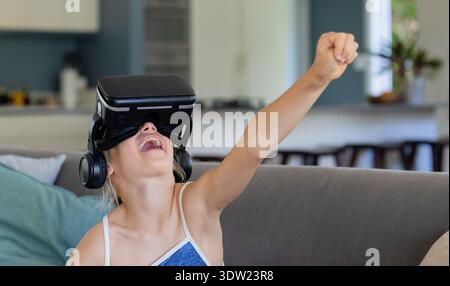 Youth female sitting on gray couch at home wearing black VR headset and headphones, laughing Stock Photo
