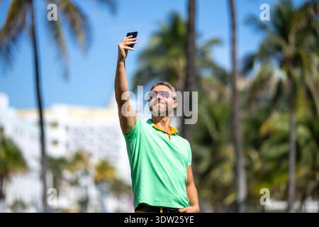 Tourist man takes a selfie near palm tree. Man captures a selfie during summer vacation. Traveler snaps a selfie in a tropical destination. A selfie Stock Photo