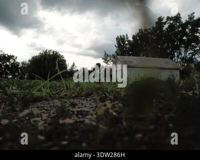 Weathered white clapboard garage or outbuilding viewed from ground level with overgrown vegetation in foreground and dramatic storm clouds overhead, H Stock Photo