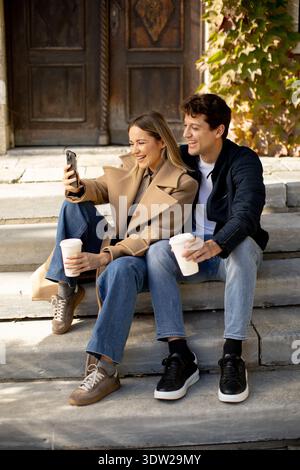 A couple sits on stone steps outside a building. They hold cups of coffee and look at a phone together. Leaves change color in the background, hinting Stock Photo
