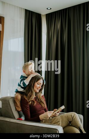 A mother sits on a sofa reading a book while her son is playfully resting on her shoulders in a living room. Sunlight shines through the curtains crea Stock Photo