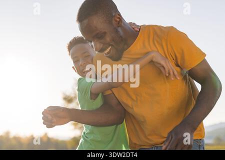 Father and son laugh while playing together outside during sunset hours. They share happy moments in nature and bond as a family. Stock Photo