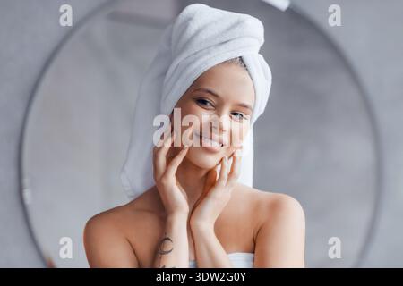 Woman With Towel on Head Smiles While Touching Her Face in a Bathroom Setting Stock Photo