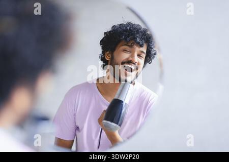Young Man Styles Hair With Hair Dryer While Smiling in Mirror at Home in Morning Stock Photo