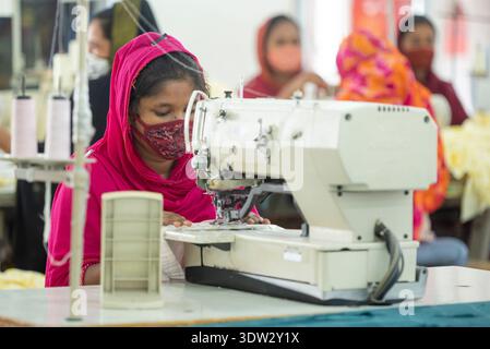 Young woman wearing pink hijab and face mask operating industrial sewing machine in busy textile factory with other workers in background. Stock Photo