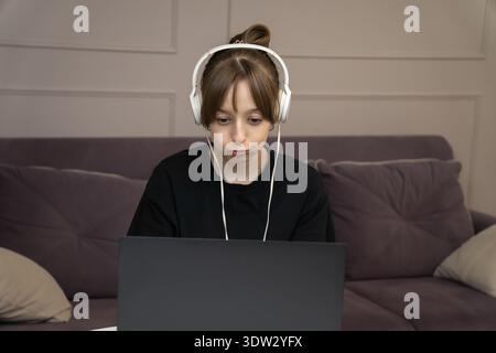 Girl wearing headphones puffing cheeks while using laptop at home, perhaps expressing boredom or frustration during online class or homework Stock Photo