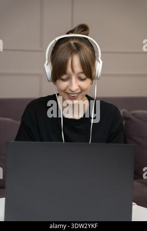 Smiling young woman using headphones at laptop, expressing joy and excitement during video call or online conference, celebrating success in home offi Stock Photo