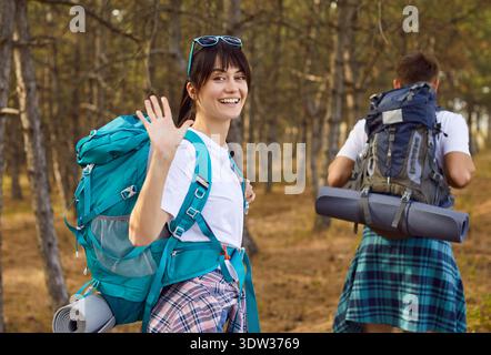 Smiling female hiker with backpack waving while partner walking on forest trail Stock Photo
