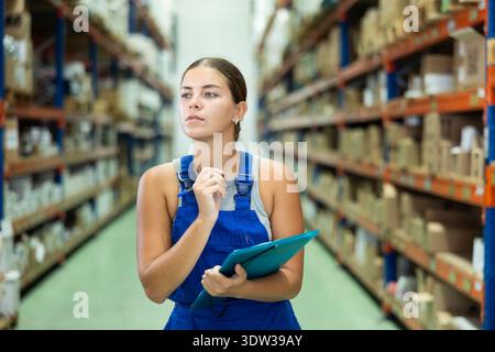 Attentive female manager keeps records of construction materials in warehouse, writing down data on paper Stock Photo