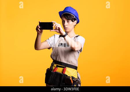 Adult female builder in safety helmet captures photo using smartphone with focused attention. Caucasian builder in construction attire taking picture and visually inspecting renovation site. Stock Photo