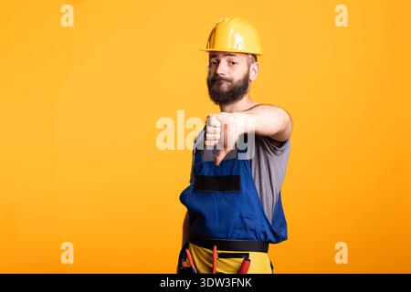 Disappointed male contractor signals disapproval with thumbs down. Man wearing protective gear stands in studio, ideal for concepts of poor results, rejection, low ratings or construction setbacks. Stock Photo