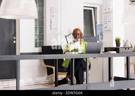 Black businesswoman seated at desk appearing confident and prepared, showing leadership potential in corporate workplace. Skilled female accountant reading and organizing company reports. Stock Photo