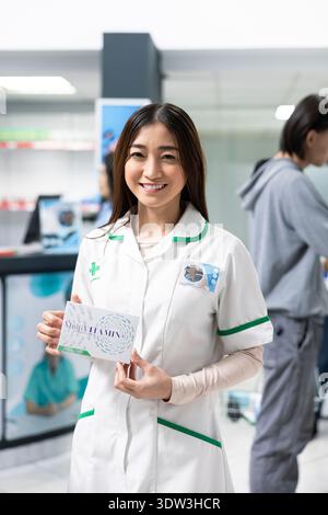 Smiling asian pharmacist holds medicine box, showcasing pharmaceutical products available in modern drugstore. Portrait of professional and approachable female drugstore attendant. Stock Photo