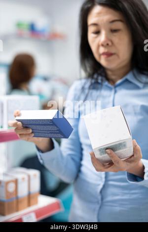 Closeup of middle aged woman comparing capsules and pills while shopping in pharmacy aisle. Focused on reading labels, female client carefully selects medications to support health and wellbeing. Stock Photo