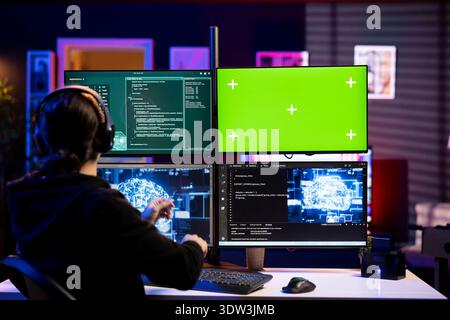 Hacker listening music using AI on green screen PC to steal credit card numbers and infiltrate banking systems. Cybercriminal wearing headphones running malicious code on chroma key computer Stock Photo