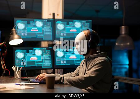 Adult man hides identity behind white mask and attempts digital intrusion involving sensitive information and network access. Hacker in hoodie and headphones hacking and retrieving encrypted data. Stock Photo