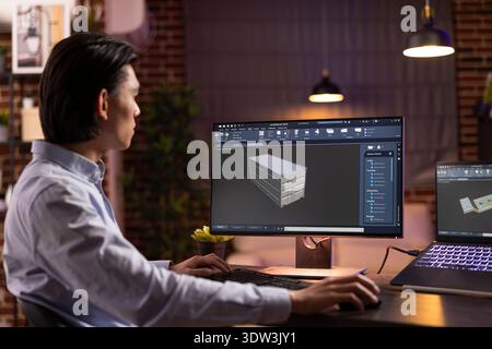 Male freelancer focusing on CAD software, creating precise 3D model structures from apartment during evening hours. Young asian man developing online industrial tools using computer and laptop setup. Stock Photo