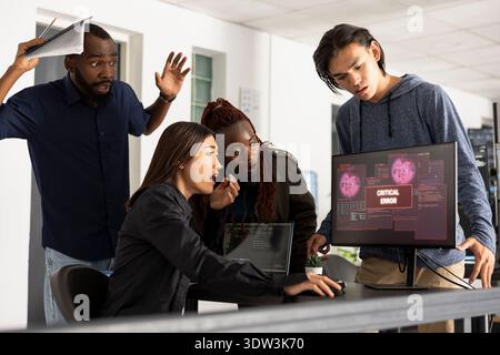 Diverse colleagues show frustration and focus while analyzing system crash on computer screen in tech startup workspace. Agile development team collaborates to solve complex programming bug. Stock Photo