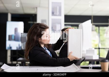 Female architect holding miniature building model and discussing project details with contractors on video call. Creative designer participates in virtual meeting, collaborating with remote client. Stock Photo