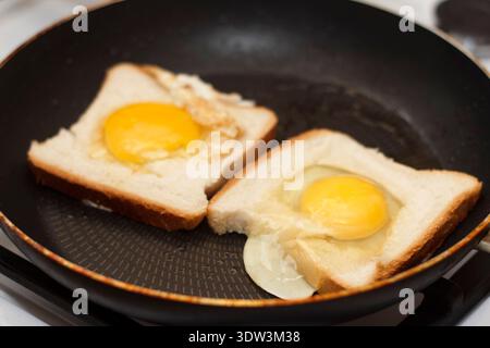 Close-up of two eggs in a basket, also known as toad in the hole, frying in a black non-stick pan. Raw yellow yolks sit in the center of white bread. Stock Photo