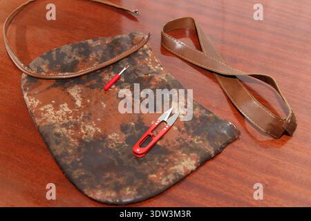 A worn, brown patterned bag is dismantled for repair on a wooden table. Red-handled thread snips and a seam ripper lay on top, ready for sewing or upc Stock Photo