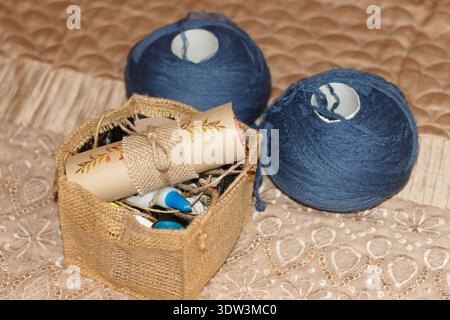 A cozy still life of crafting supplies. Two balls of blue yarn and a burlap basket with a decorated scroll sit on a quilted beige fabric. Warm, natura Stock Photo