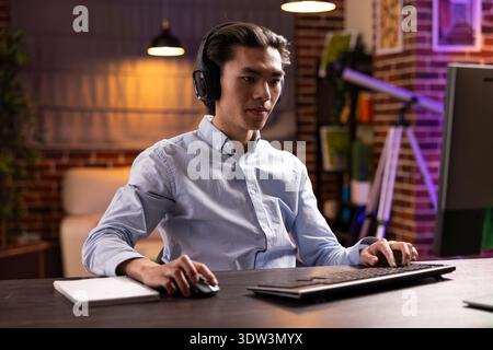 Young asian man with headphones concentrating on pc screen, watching professional webinar. Male remote worker participating in online training, studying business strategies in home office. Stock Photo