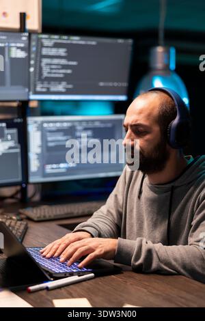 Male adult freelancer types on laptop in tech workspace, creating software while planning and debugging encrypted programs. White man works night hours on cybersecurity, analyzing spyware risks. Stock Photo
