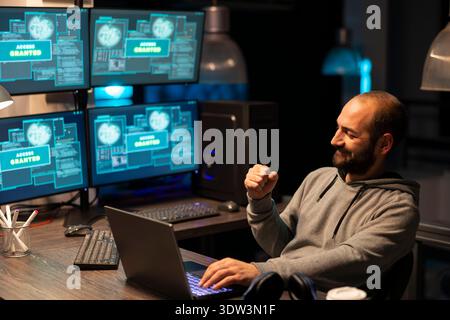 Smiling male programmer clenches fist in success after receiving access granted notification on device. Cheerful developer sits at desk with laptop, celebrating successful digital task completion. Stock Photo
