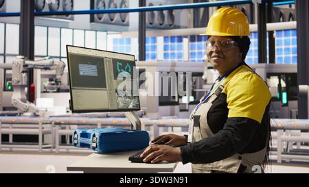 Portrait of cheerful factory engineer typing on PC, overseeing monitoring of production lines. Jolly woman in industry 4.0 plant ensuring machinery operates at optimal efficiency, camera A Stock Photo