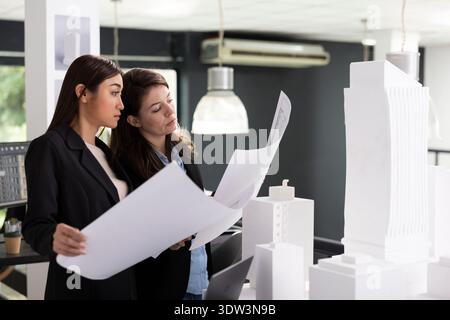Skilled architects study printed architectural blueprints, preparing for next construction site evaluation. Professional women crosschecking design variations for accurate project implementation. Stock Photo