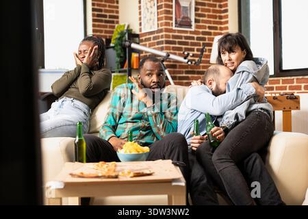 Frightened friends watch horror movie together in apartment, reacting to shocking scene with fear and nervous expressions. Caucasian couple hugs as diverse group bonds over thriller film at home. Stock Photo