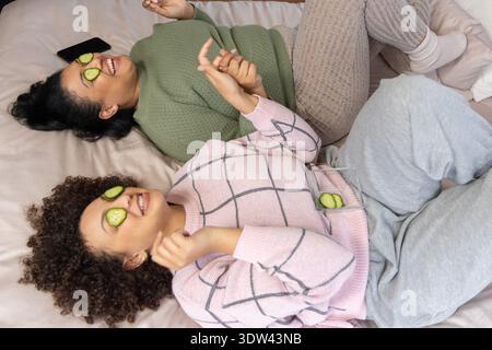 Diverse female friends lying on bed at home wearing sweaters, holding hands, using cucumber slices Stock Photo