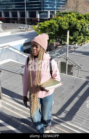 Female student walking up urban stairs wearing pink beanie hoodie holding notebooks phone backpack Stock Photo