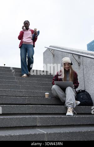 Female students walking on exterior stairs, one sitting using laptop, other talking on phone Stock Photo